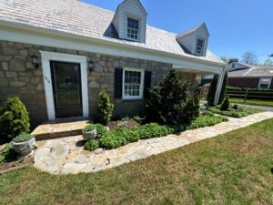 Stone house with a slate roof, two dormer windows, and a black front door, surrounded by a stone walkway and manicured shrubs.