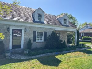 Stone house with a slate roof and two dormer windows, white trim, and black shutters in a sunny yard.