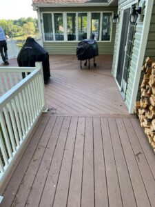 Back deck with two covered grills, sliding glass doors, and a woodpile beside the house; trees in the yard beyond the railing.