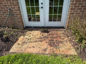 Brick exterior doorway with white-framed glass doors; garden hose hanging on left and bare, mulched planting beds nearby.