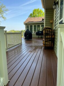 A wooden deck attached to a house with a white railing, overlooking trees and a blue sky; a covered grill and a stack of firewood are on the deck.